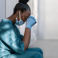Tired depressed female african nurse wearing face mask sits on hospital floor.