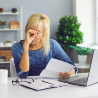 Upset senior woman sitting at desk, holding bankruptcy or debt notification from bank