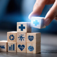 A person is stacking wooden blocks with a medical symbol on top. The blocks are arranged in a pyramid shape, with the top block featuring a heart and a cross