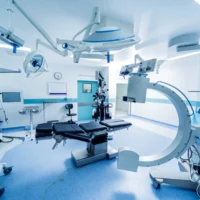 Modern medical operating room with advanced equipment including surgical lights, monitors, and a C-arm machine. The room is bright and sterile.