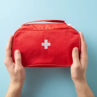 Hands hold a red first aid kit with a white cross on a light blue background, conveying preparedness and safety.